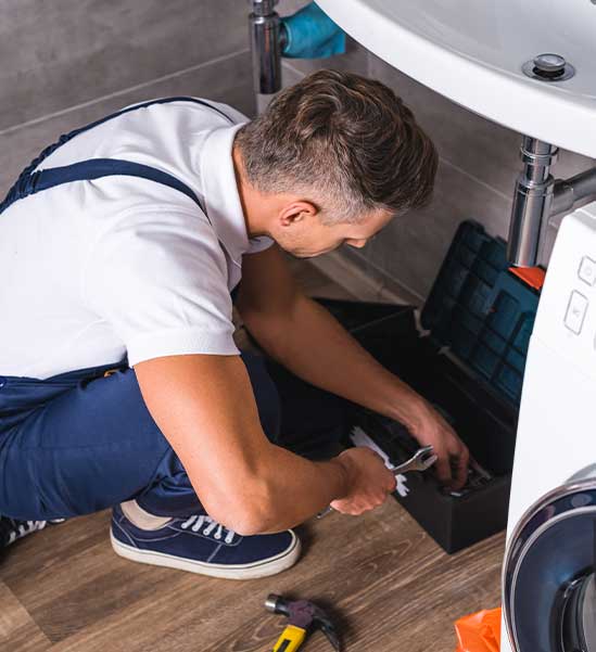 Plumber working beneath a sink beside a washing machine.